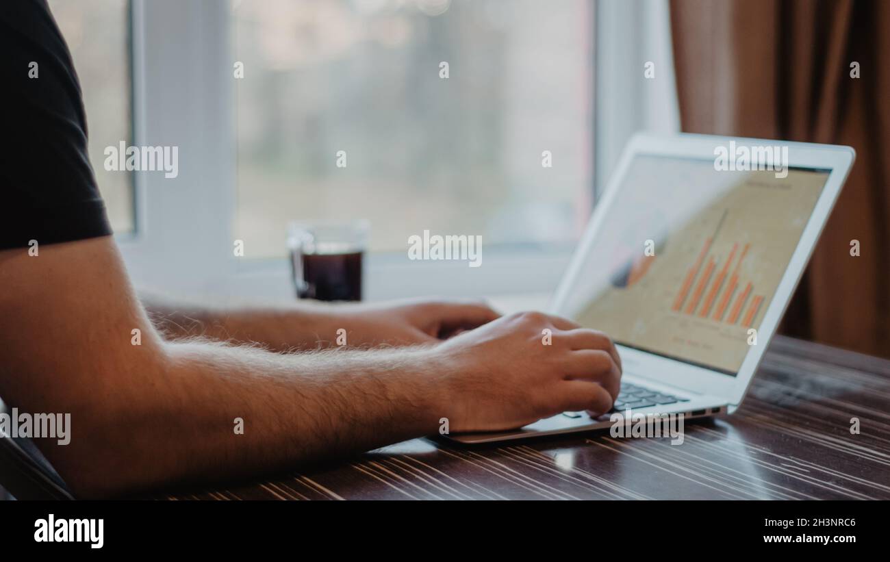 Gros plan d'un homme qui tape sur un ordinateur portable et qui travaille avec un tableau financier sur une table de bureau en bois désordonnée. Banque D'Images