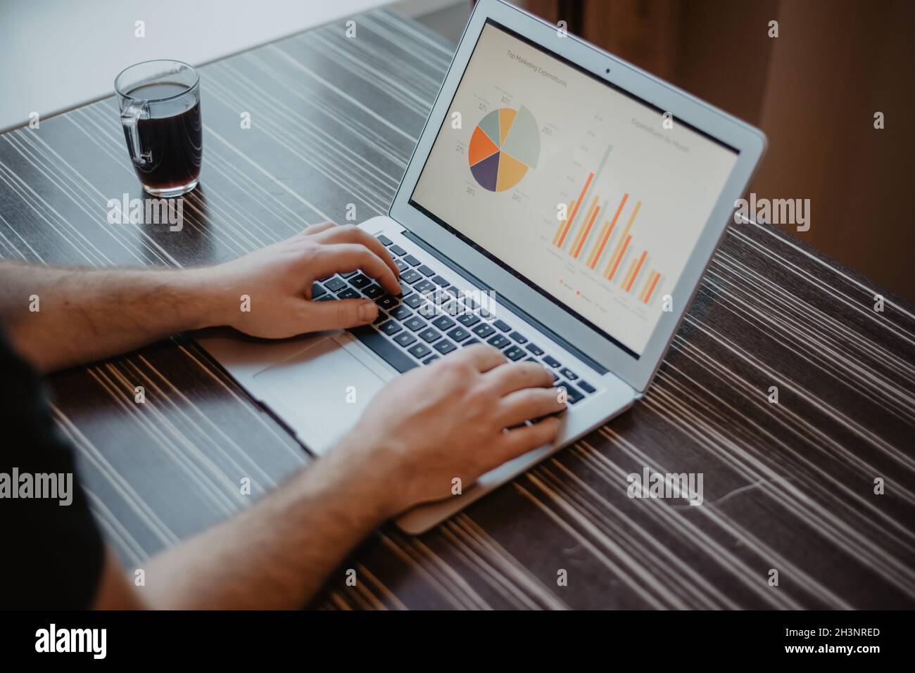 Gros plan d'un homme qui tape sur un ordinateur portable et qui travaille avec un tableau financier sur une table de bureau en bois désordonnée. Banque D'Images