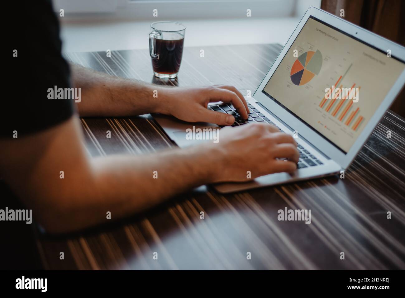 Gros plan d'un homme qui tape sur un ordinateur portable et qui travaille avec un tableau financier sur une table de bureau en bois désordonnée. Banque D'Images