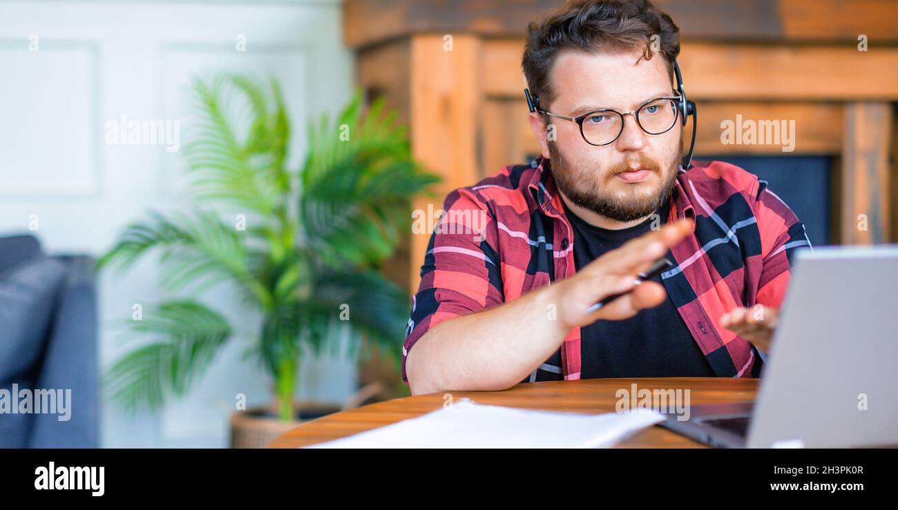 Seri8ous jeune homme en lunettes travaillant à la maison - il est assis devant l'écran d'ordinateur portable et ayant des appels vidéo en ligne.Formatez la photo 2x1. Banque D'Images