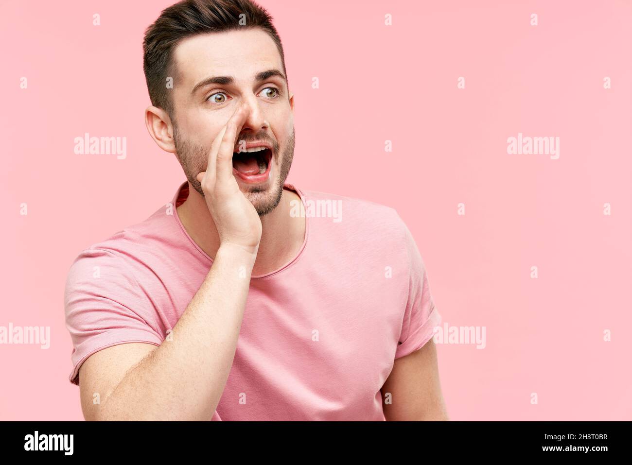 Jeune homme beau criant et criant fort à côté avec la main sur la bouche et l'espace de copie pour le texte sur fond rose Banque D'Images