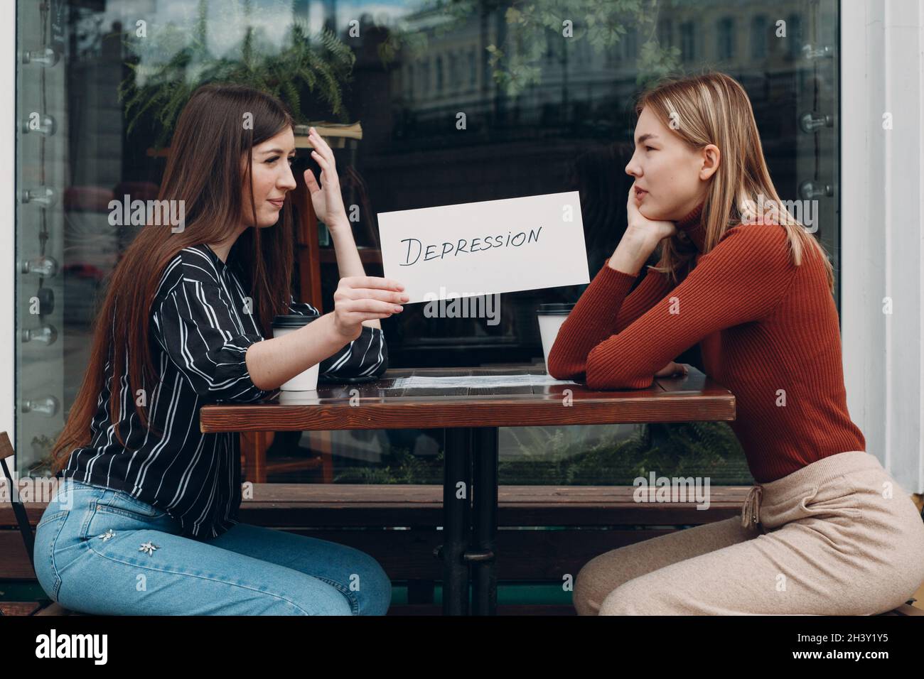 Femme tenant une feuille de papier blanc étiquetée dépression mot dans la main. Deux femmes parlent dans un café de rue. Concept de dépression cachée Banque D'Images