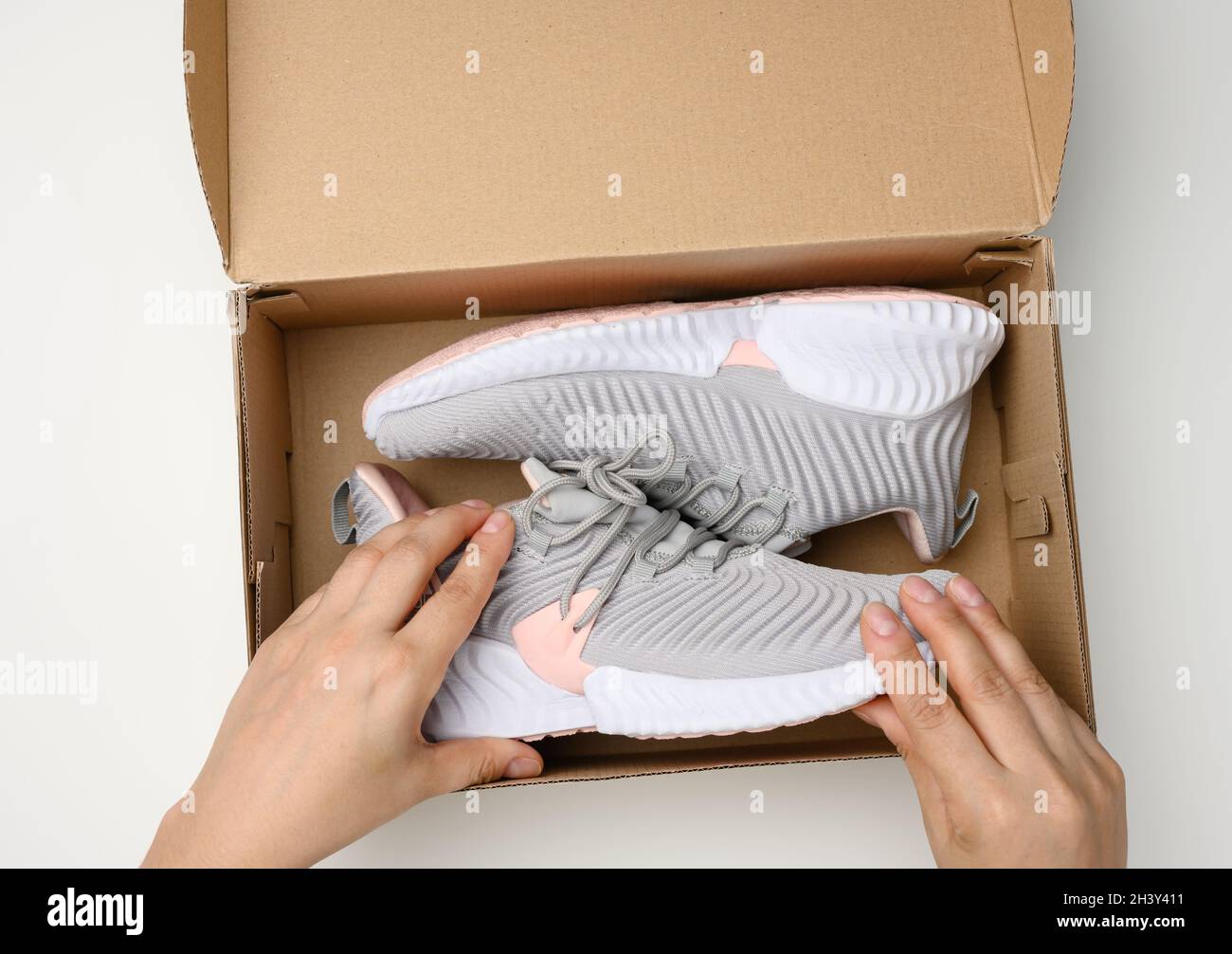 Deux mains femelles tiennent une boîte en carton marron avec des baskets de sport grises sur une table blanche, inspectant une commande en ligne Banque D'Images