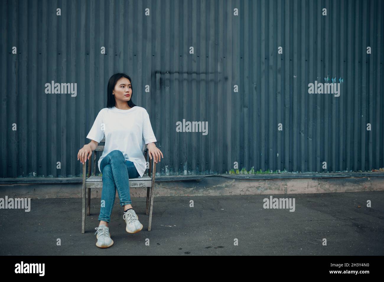 Jeune femme asiatique urbaine assise sur une chaise près du mur bleu sur la rue à l'extérieur. Banque D'Images