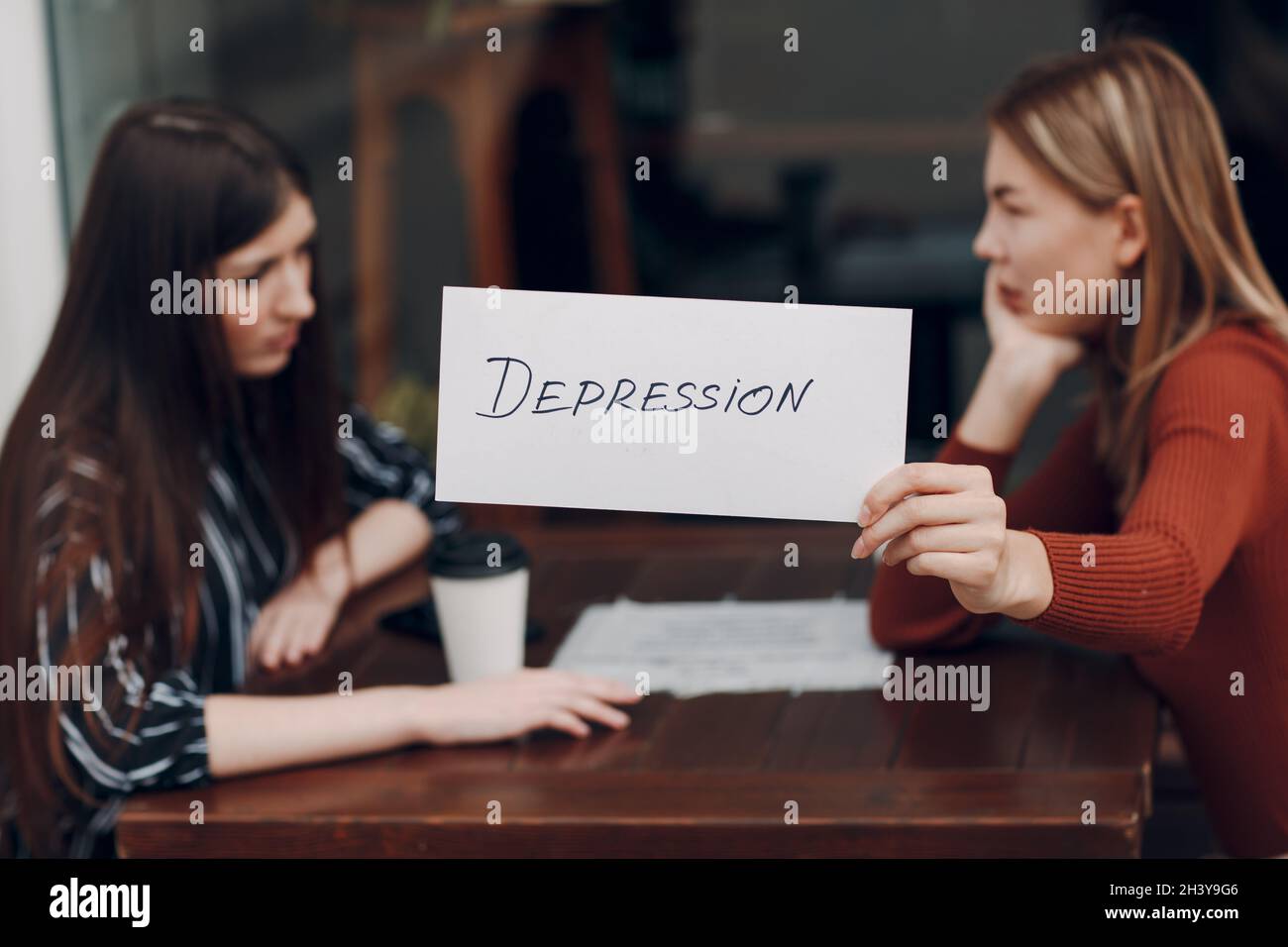Femme tenant une feuille de papier blanc étiquetée dépression mot dans la main. Deux femmes parlent dans un café de rue. Concept de dépression cachée Banque D'Images