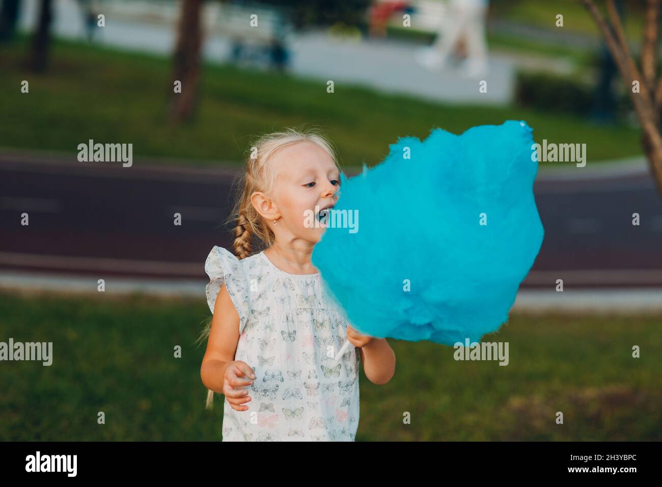 Petite fille blonde mangeant des bonbons en coton bleu dans le parc. Banque D'Images