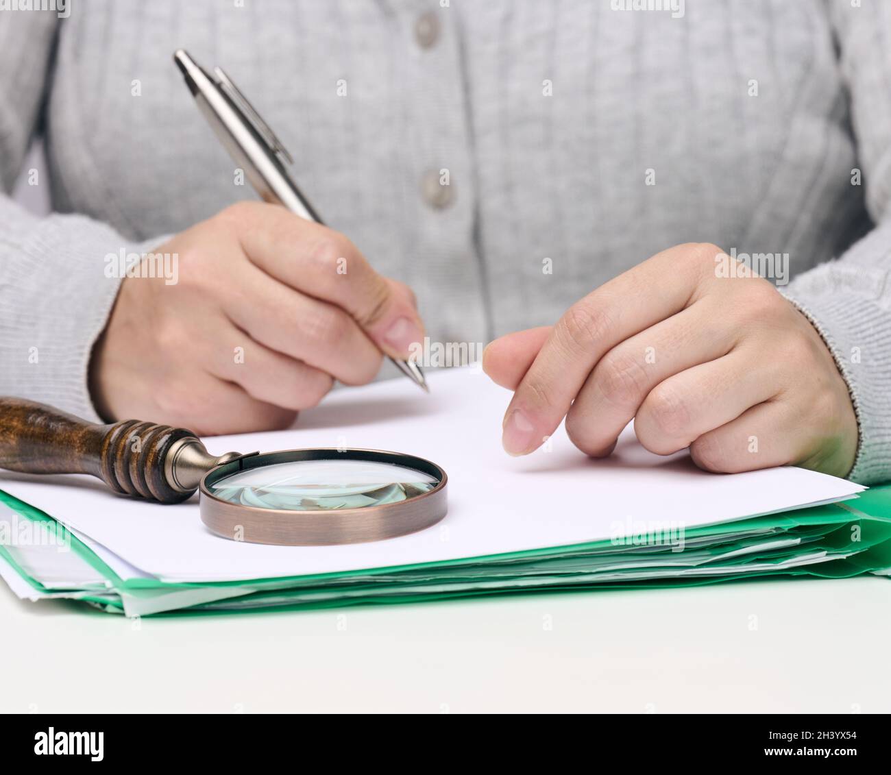 Femme s'assoit à une table blanche et tient un stylo en métal sur une pile de papiers, à l'autre main une loupe Banque D'Images