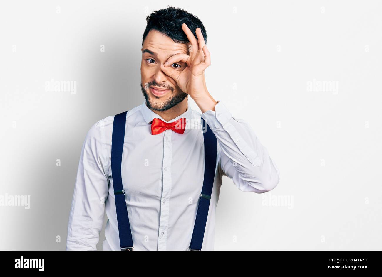 Homme hispanique avec barbe portant un look hipster avec noeud papillon et  bretelles faisant un geste ok avec la main souriante, oeil regardant à  travers les doigts avec fa heureux Photo Stock -