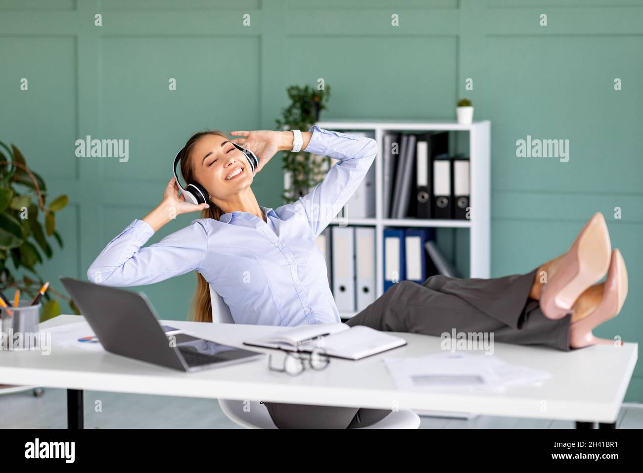 La vie est bonne.Une femme entrepreneure joyeuse écoute de la musique sur le lieu de travail et met des pieds sur la table Banque D'Images