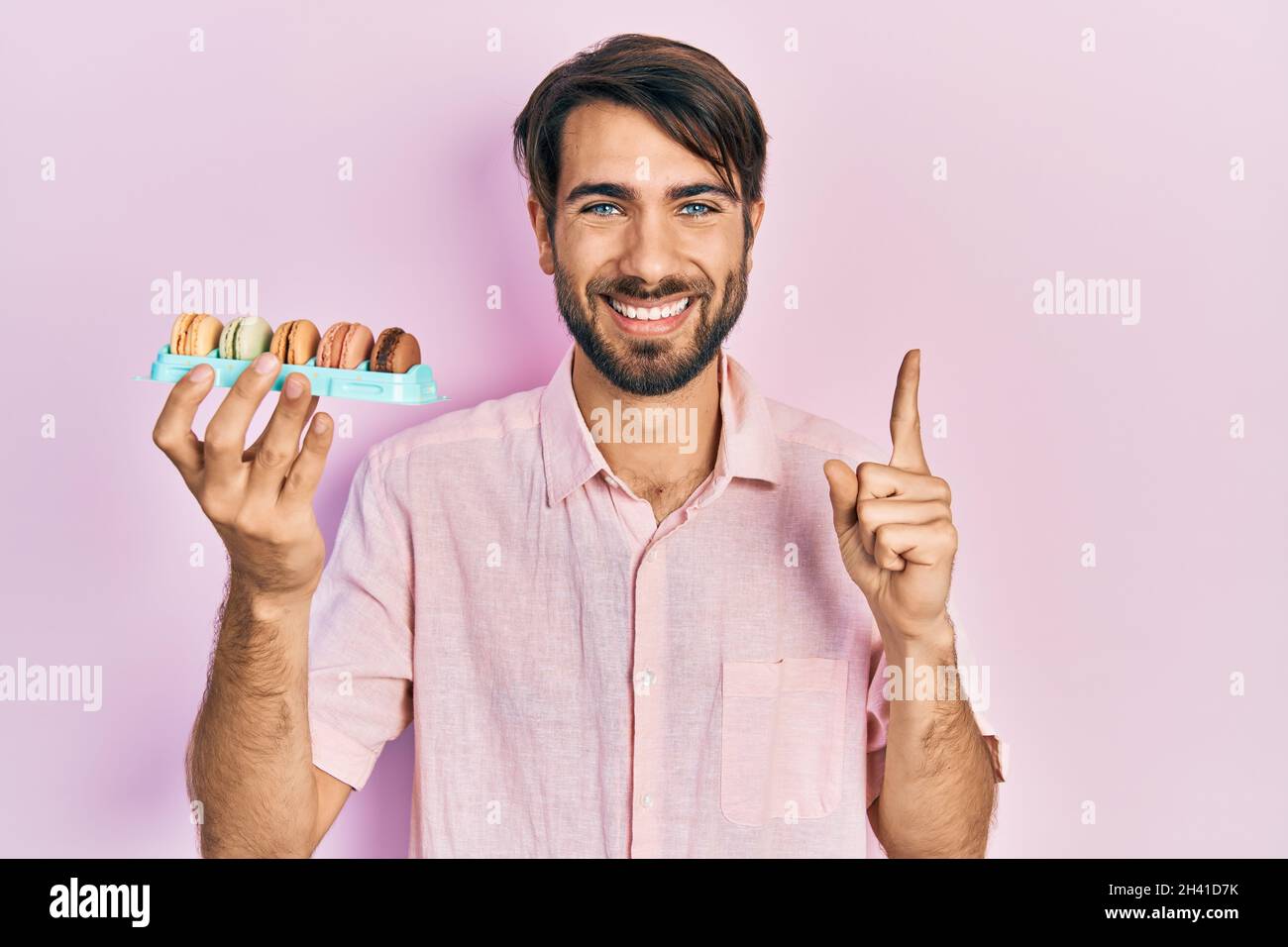 Jeune homme hispanique portant de délicieux macrons pâtisseries souriant avec une idée ou une question pointant du doigt avec le visage heureux, numéro un Banque D'Images
