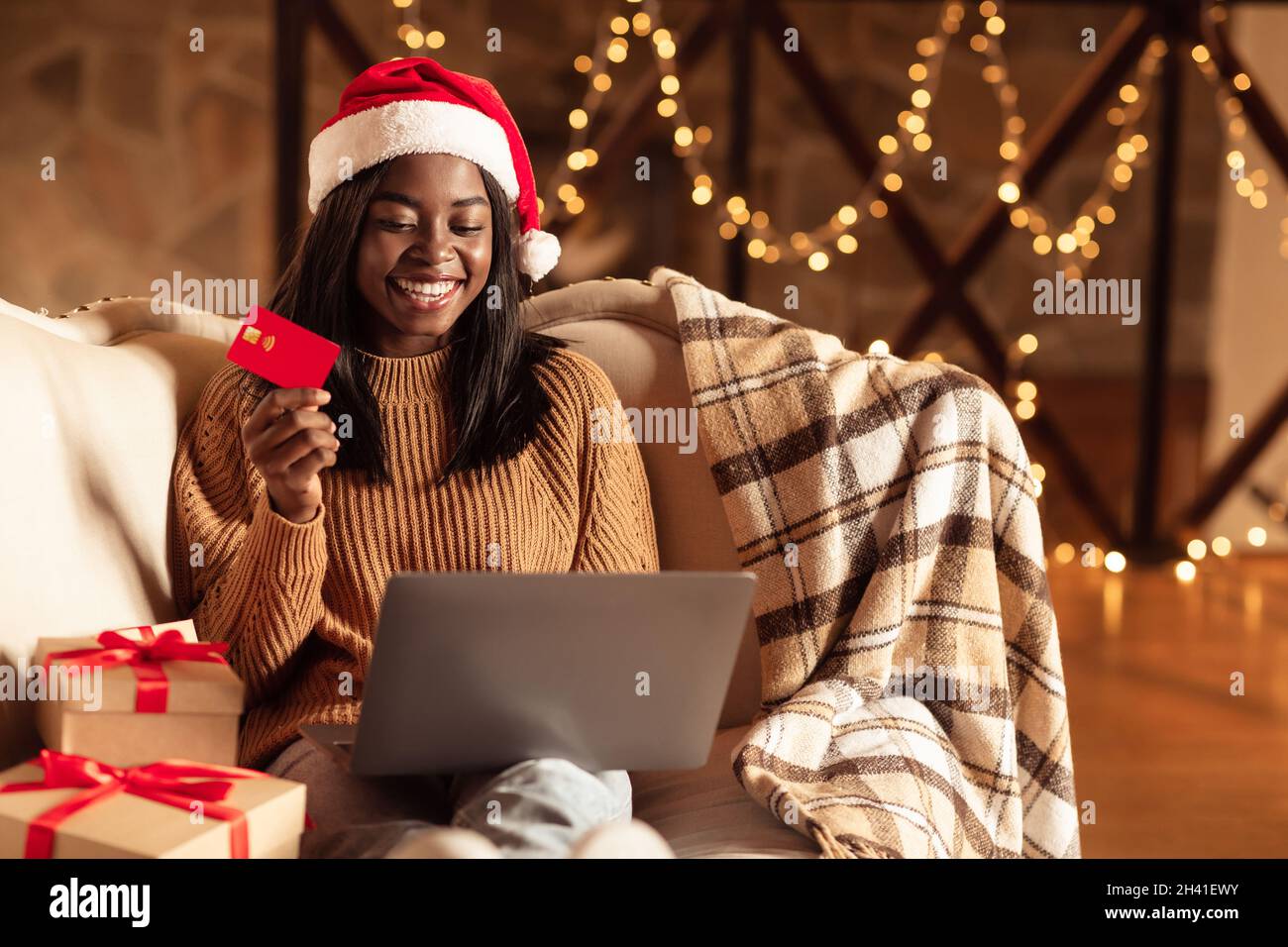 Achats de Noël en ligne.Une femme noire dans un chapeau de père Noël assis  près des boîtes-cadeaux, tenant une carte de crédit et utilisant un  ordinateur portable à la maison Photo Stock -