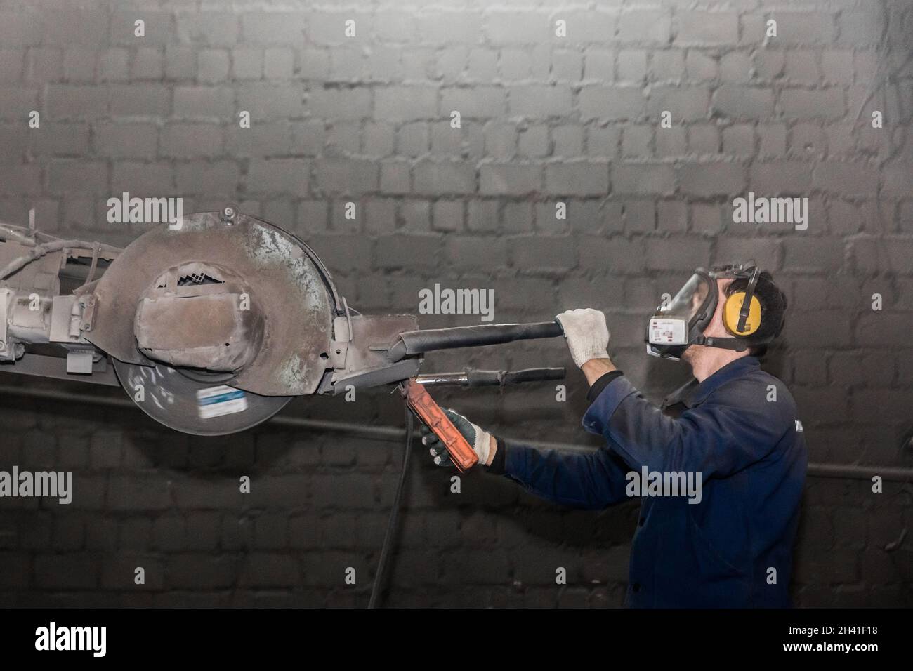 Un homme en vêtements de travail et un respirateur, contrôle l'équipement lourd avec pierre à affûter pour nettoyer les structures en béton armé dans un atelier d'une industrie Banque D'Images
