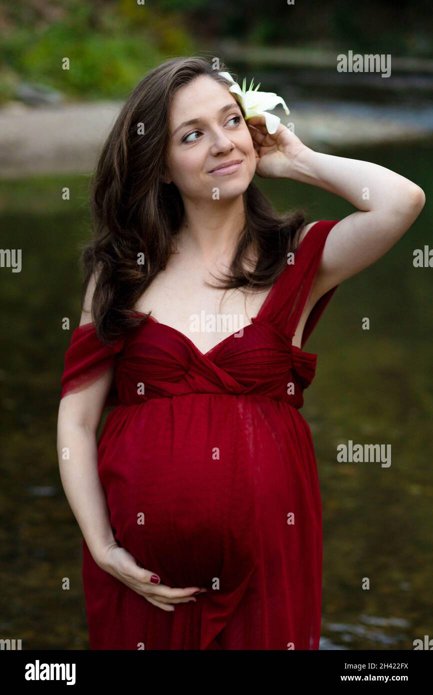 Jeune femme enceinte blonde, élégante, debout dans la rivière en robe rouge, fleur blanche derrière son oreille Banque D'Images