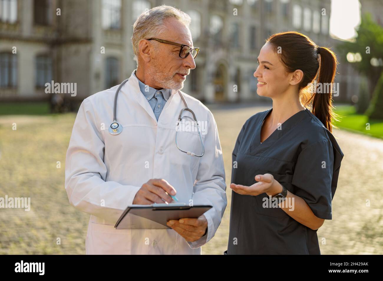 Portrait d'un médecin de laboratoire en blouse et en lunettes tenant le presse-papiers, en regardant une jeune infirmière souriante, debout ensemble à l'extérieur près de la clinique Banque D'Images