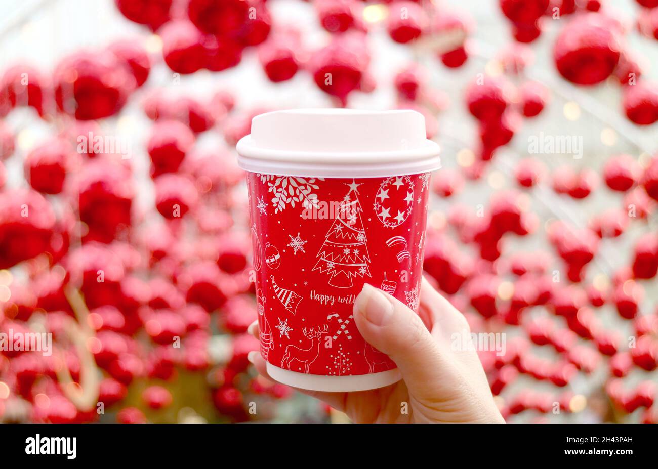 Main de femme tenant une tasse à café à thème de vacances avec des ornements boule rouge floue en arrière-plan Banque D'Images