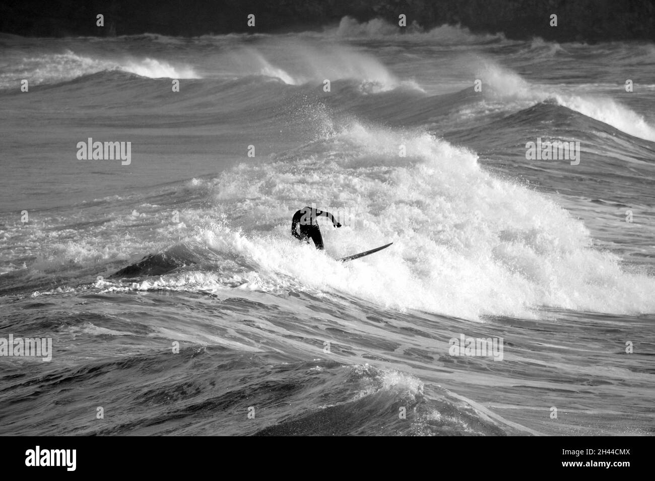 Un surfeur surfant bravement sur les vagues d'hiver près de Huntcliff, Saltburn-by-the-Sea, North Yorkshire. Photographié depuis l'embarcadère de Saltburn. Noir et blanc. Banque D'Images