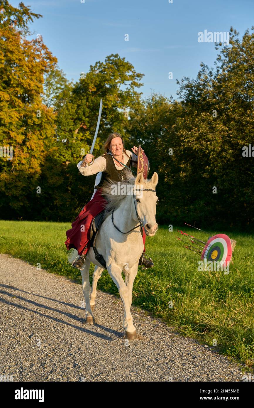 Rider féminin en costume médiéval avec épée relevée et tresses de bouclier sur le White Horse sur Un chemin naturel.Arrière-plan Green Meadow, arbres d'automne, ciel bleu. Banque D'Images