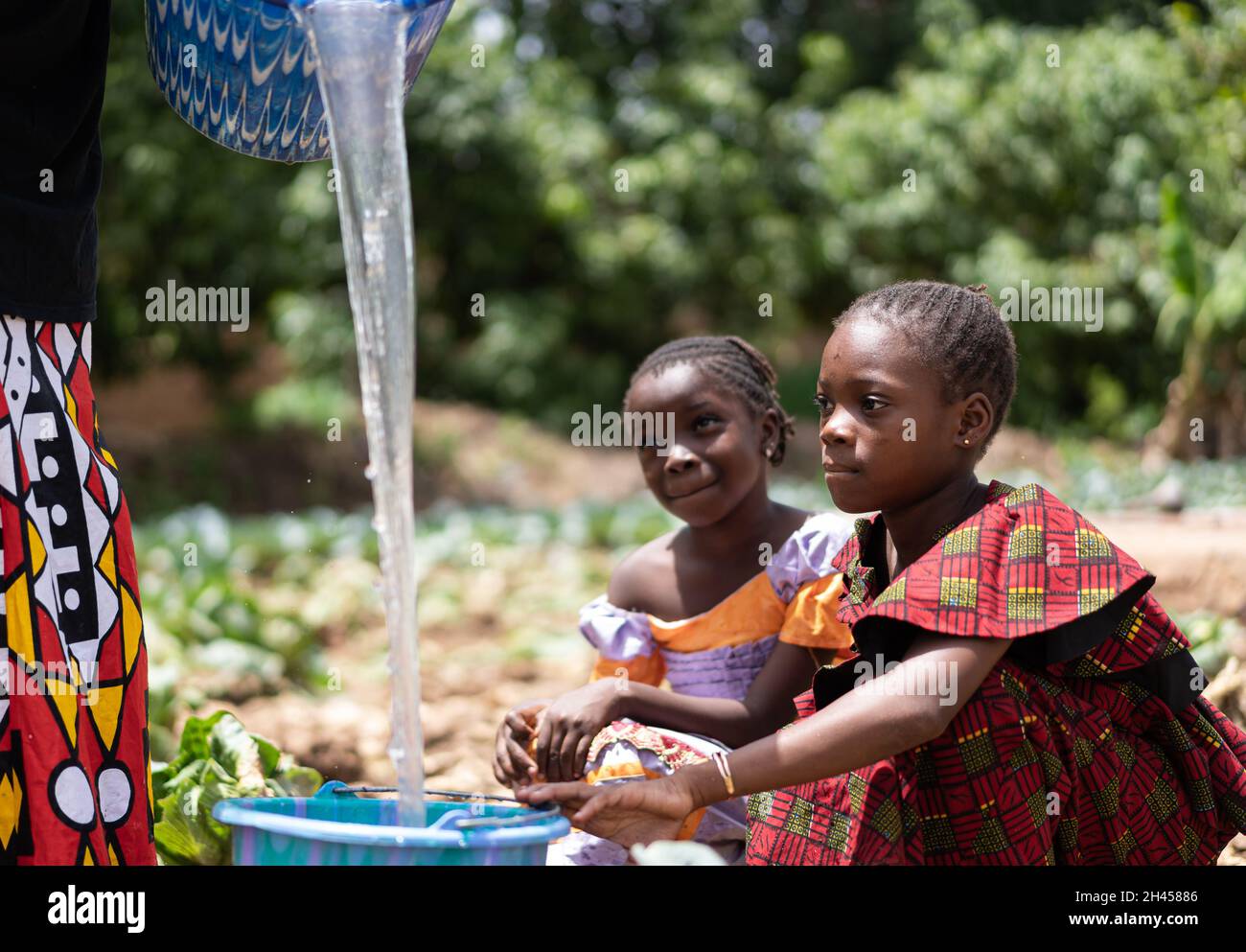 Deux petites filles noires africaines assises dans un champ et regardant un jet d'eau claire précieuse versé dans un seau ; concept de pénurie d'eau Banque D'Images