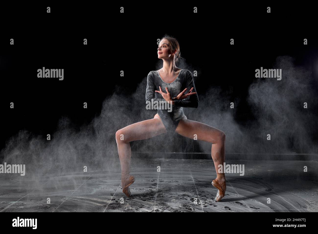 Femme flexible dansant avec de la farine sur fond noir en studio, se déplaçant avec les jambes séparées. Danseur de ballet d'athlète talentueux en combinaison au ralenti, lo Banque D'Images