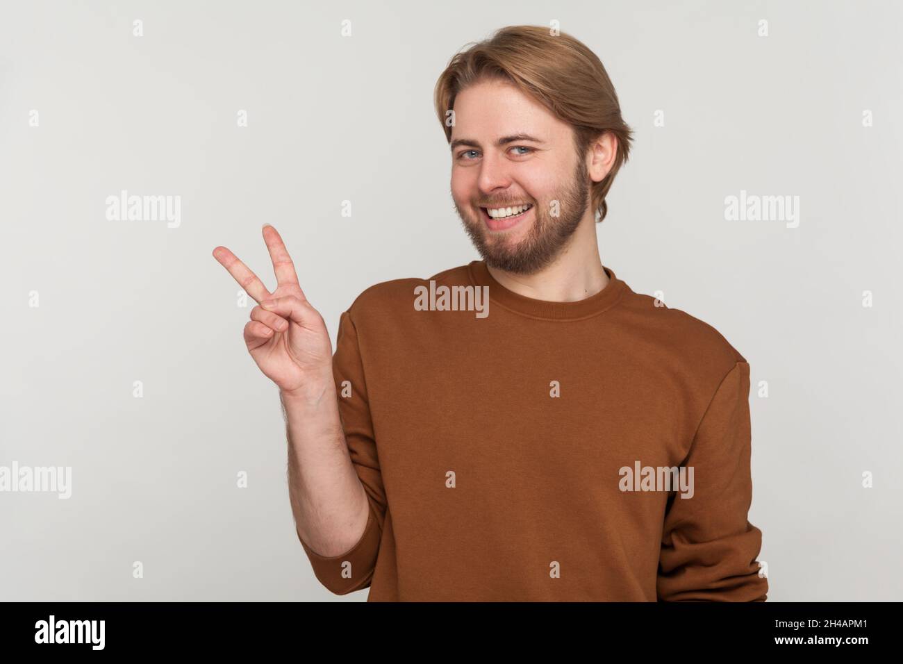 Portrait de l'homme avec la barbe portant un sweat-shirt, faisant le geste de victoire et souriant à l'appareil photo, montrant la paix, le signe v avec les deux doigts.Prise de vue en studio isolée sur fond gris. Banque D'Images