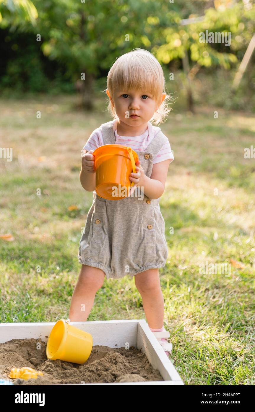 Une petite fille sérieuse dans une combinaison grise avec short avec un seau de jouet orange avec le sable dans ses mains joue dans le jardin.Il y a d'autres jouets Banque D'Images