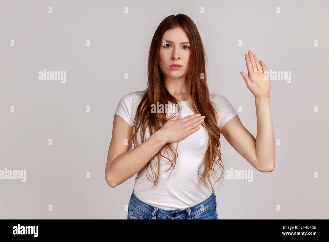 Portrait d'une femme très responsable et honnête donnant la promesse, faisant solennellement vœu dans la tradition cérémoniale avec la main levée, portant un T-shirt blanc.Prise de vue en studio isolée sur fond gris. Banque D'Images