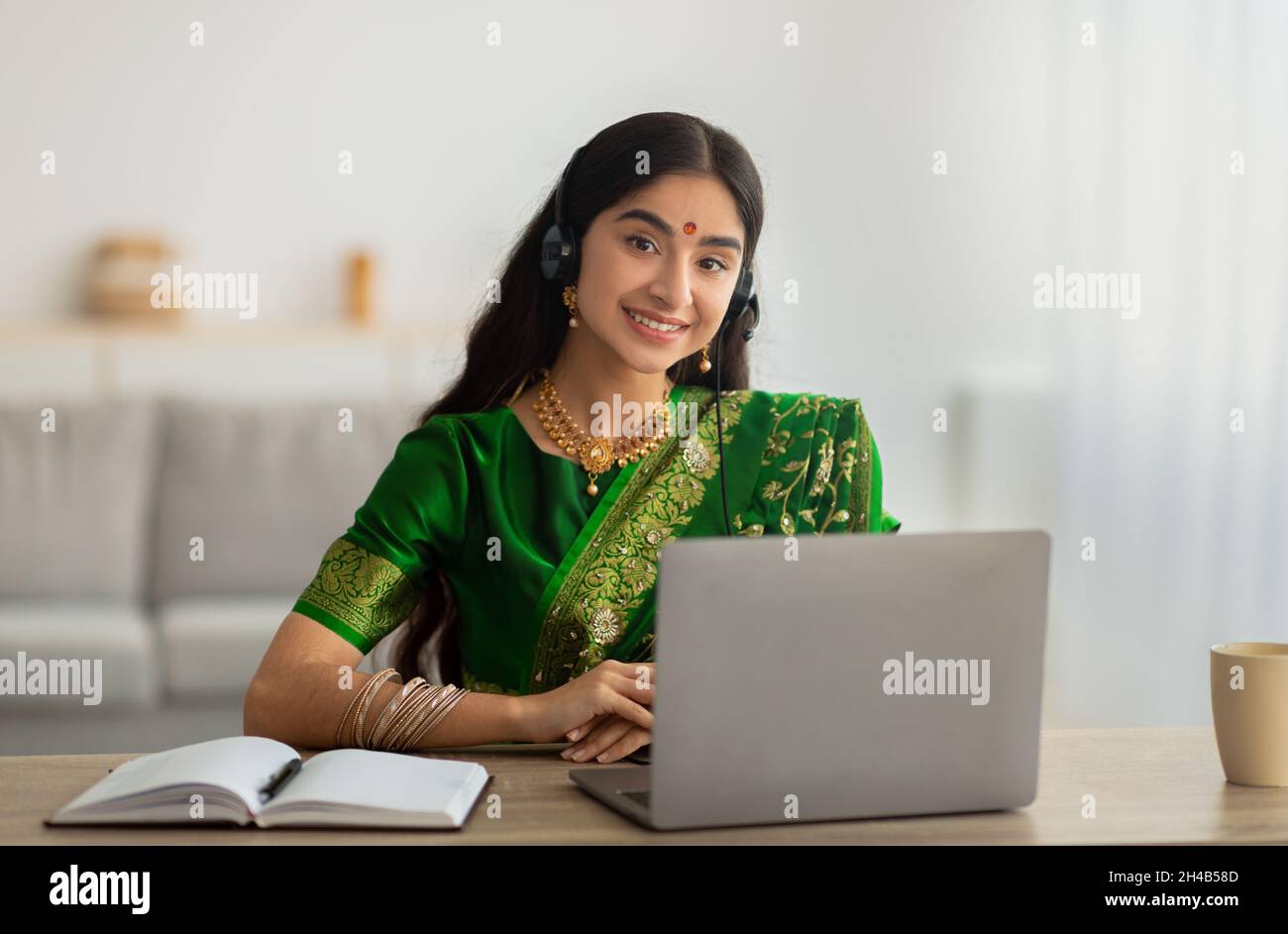 Portrait d'une femme indienne gaie en robe sari portant un casque, ayant une réunion en ligne ou une conférence Web sur ordinateur portable à la maison.Une étudiante prend p Banque D'Images