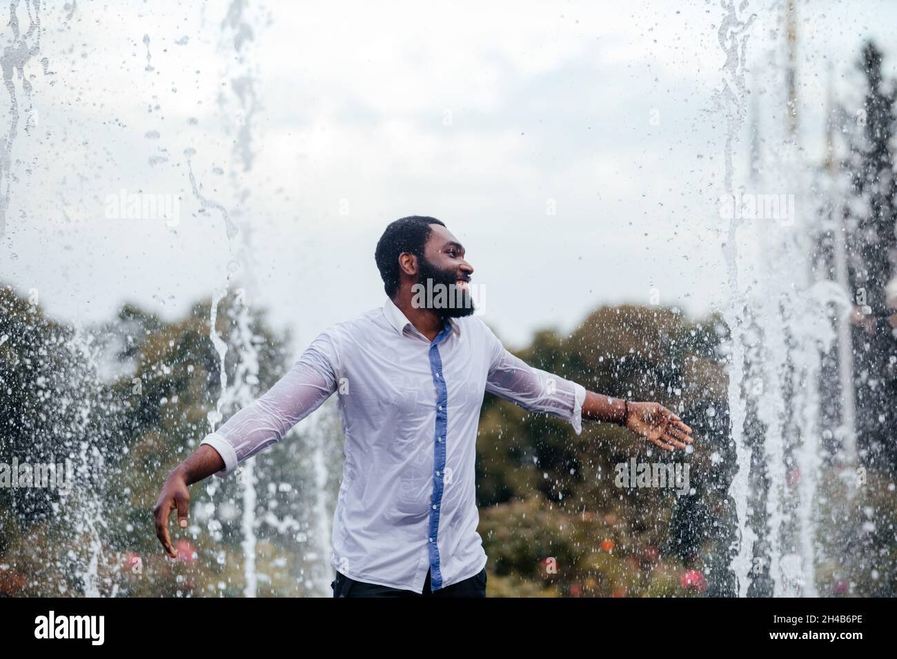 Le jeune homme souriant s'amuse dans une fontaine de la ville.Il porte une chemise blanche mouillée Banque D'Images