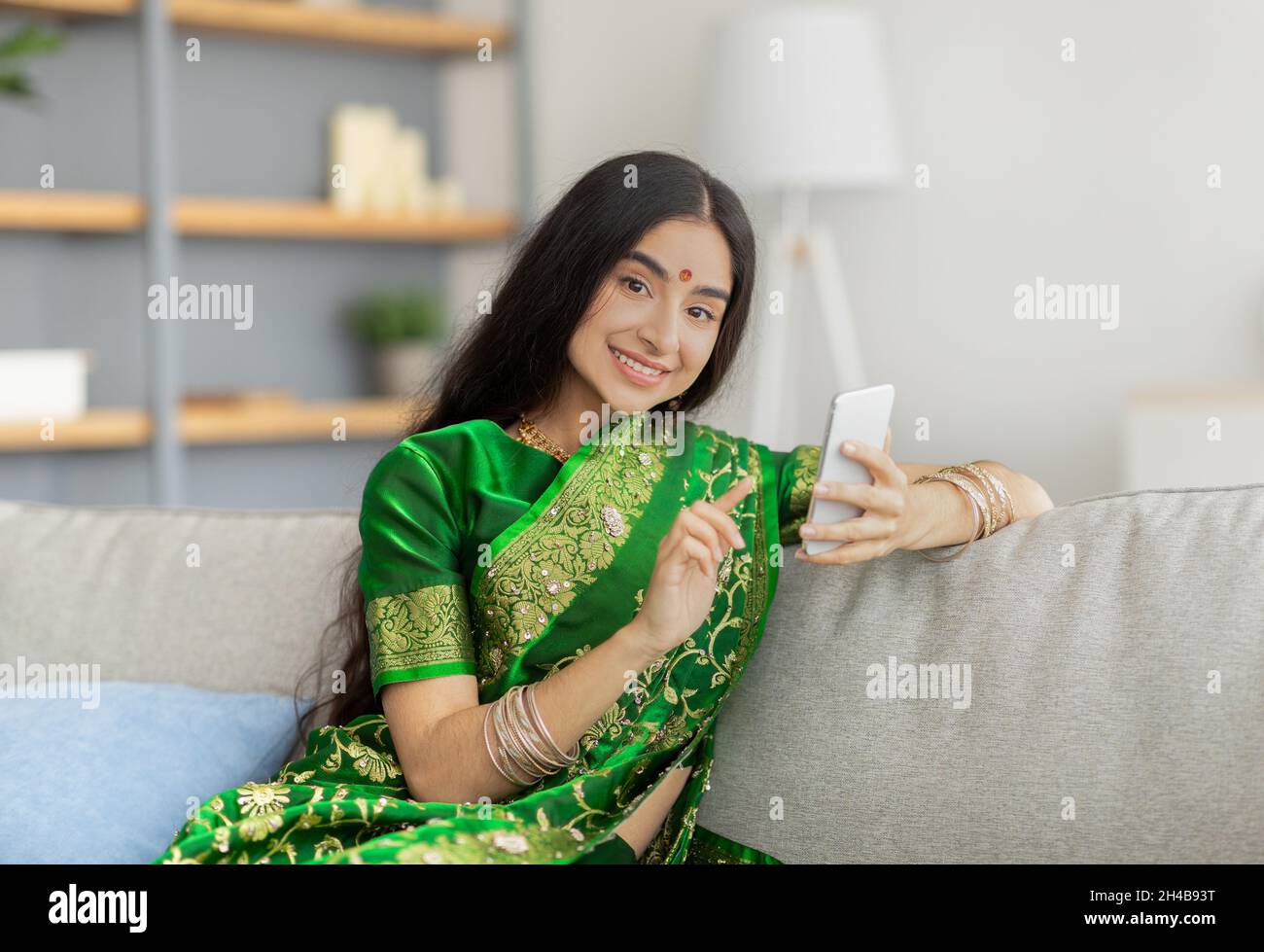 Portrait de la belle femme indienne en robe sari en utilisant le téléphone portable sur le canapé à la maison.Charmante jeune femme orientale dans des vêtements ethniques à la recherche de web, échec Banque D'Images