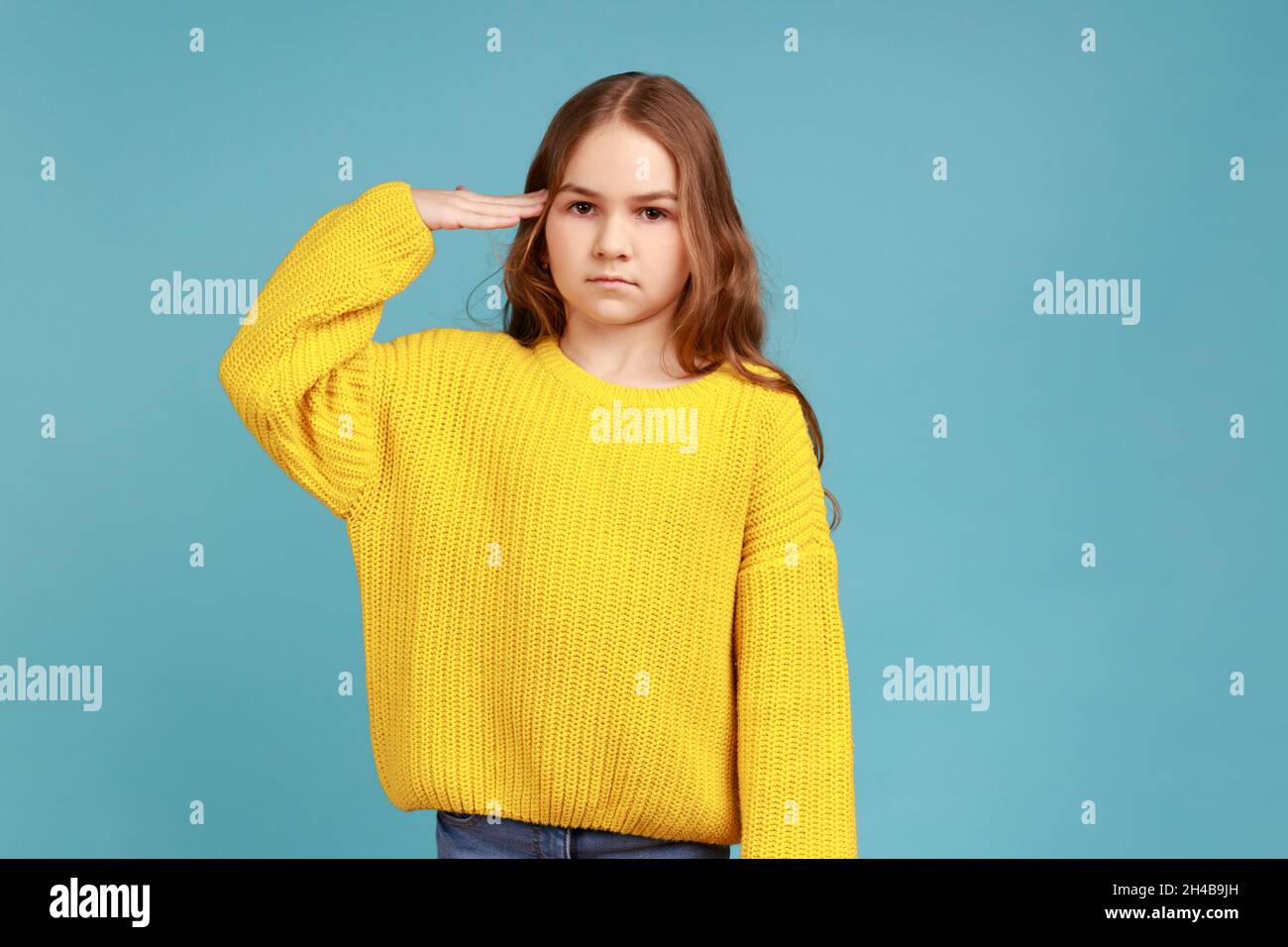 Oui, monsieur.Portrait de petite fille saluant et regardant l'appareil photo avec respect, enfant patriotique, portant jaune décontracté style chandail.Studio d'intérieur isolé sur fond bleu. Banque D'Images