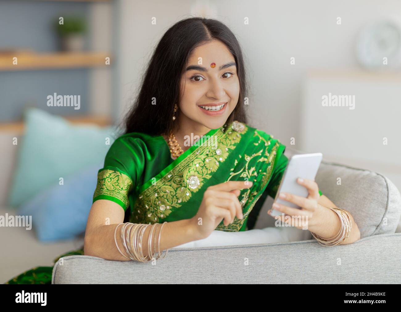 Charmante dame indienne dans une maison de saree traditionnelle assise sur un canapé avec téléphone portable, faisant appel vidéo à un ami de la maison.Femme orientale millénaire en cl ethnique Banque D'Images