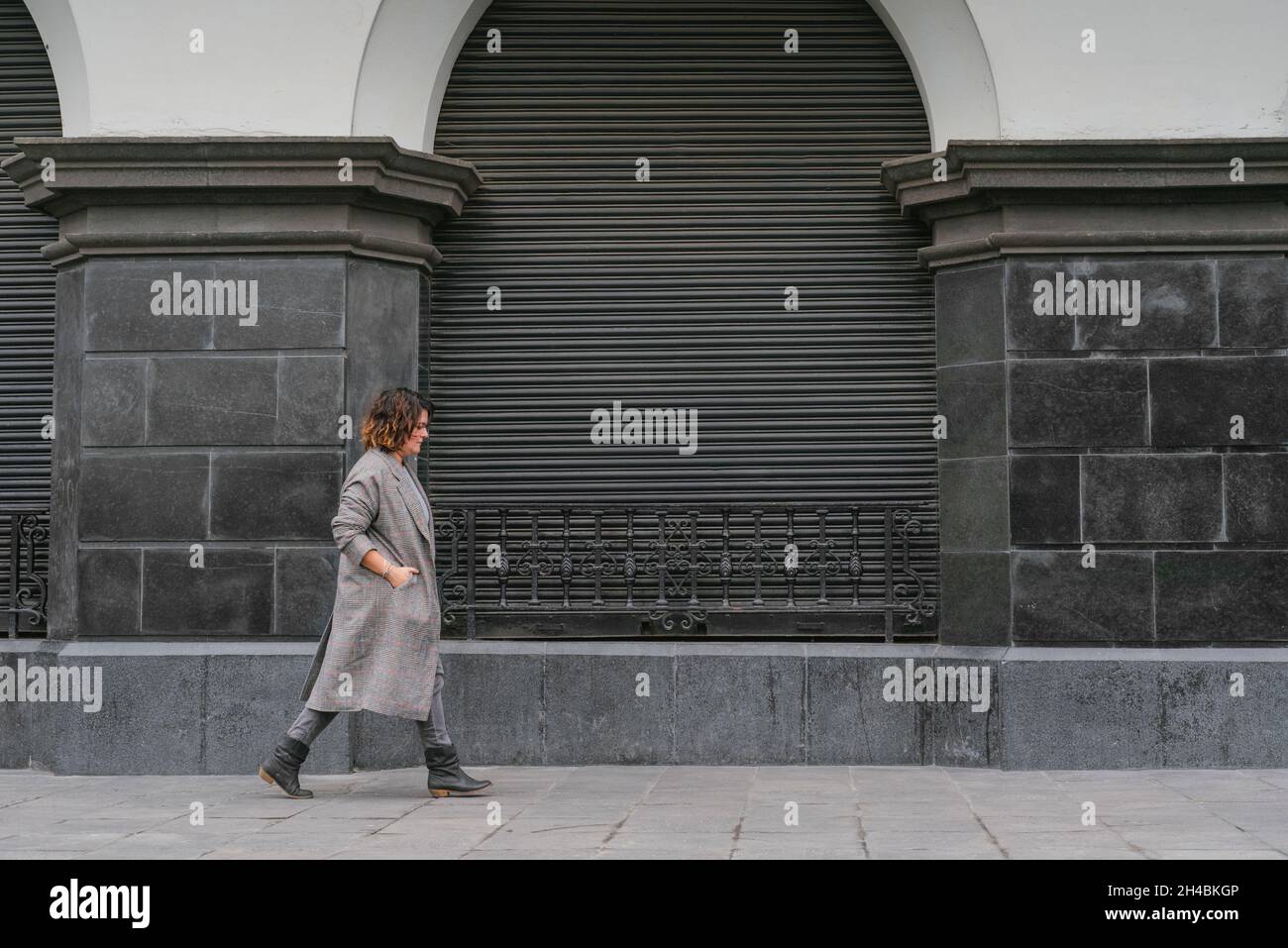 Une femme descend une vieille rue en regardant le sol et a ses mains à l'intérieur de son long manteau, sur un fond d'architecture en niveaux de gris anciens. Banque D'Images