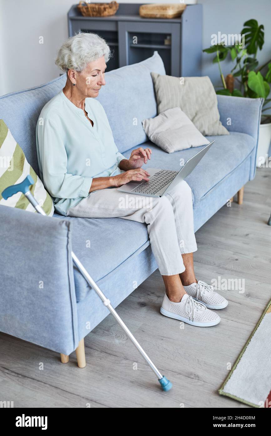 Portrait vertical complet d'une femme âgée aux cheveux blancs utilisant un ordinateur portable tout en se relaxant sur un canapé à la maison Banque D'Images