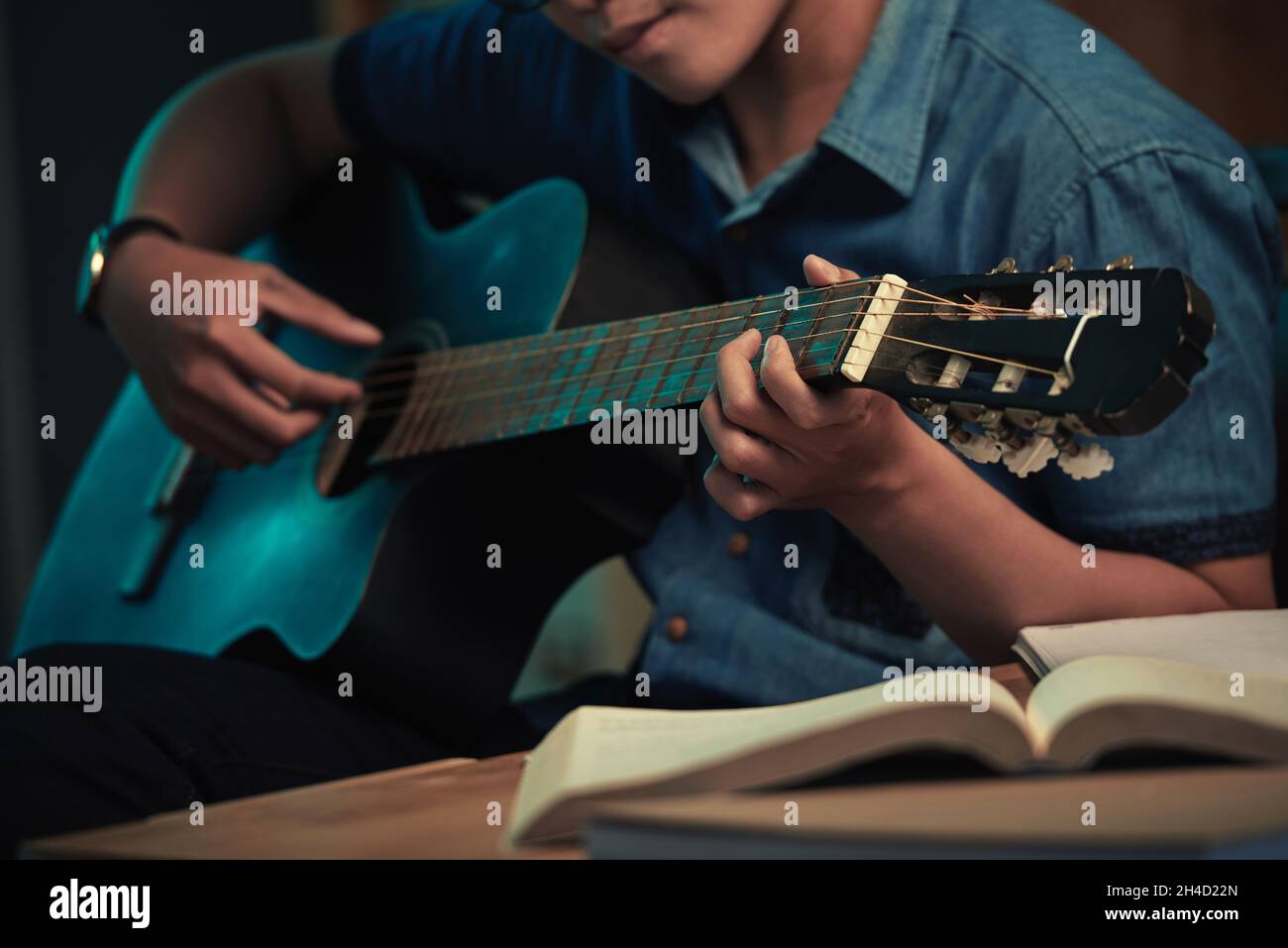 Gros plan d'un jeune homme dans une chemise décontractée assis à table avec un livre ouvert et jouant de la guitare dans l'obscurité Banque D'Images
