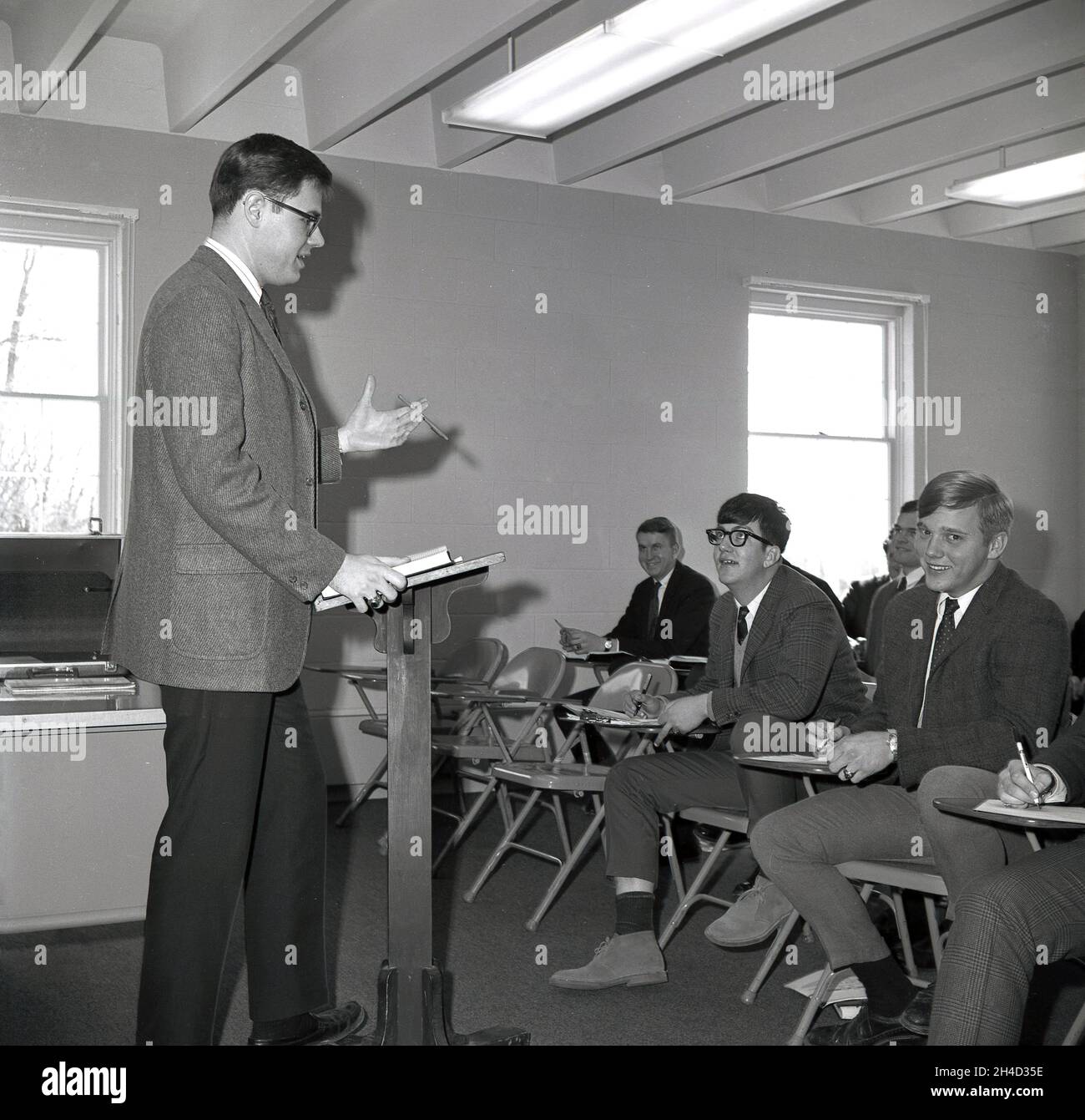 Années 1960, des étudiants de haut niveau historiques et bien habillés d'un collège d'affaires assis dans une pièce sur des chaises en métal avec de petits bureaux latéraux, à l'écoute d'un enseignant de sexe masculin qui est debout dans un pupitre en bois, Monroe College, Middletown, Virginie, États-Unis. Banque D'Images