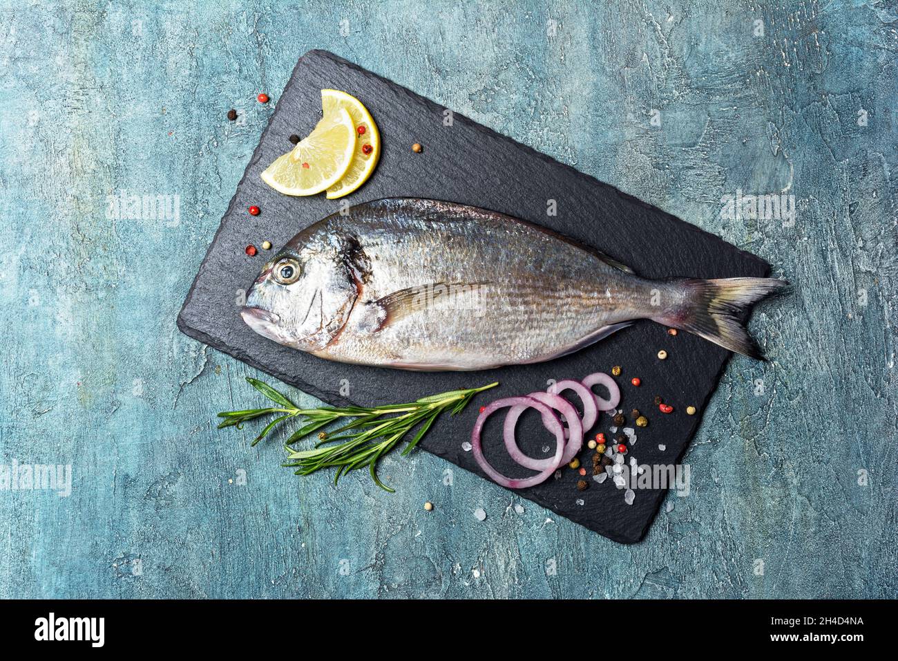 Vue de dessus de poisson frais dorado ou dorade à tête dorée avec citron, oignon et épices comme poivre, romarin, sel sur ardoise noire et fond de béton bleu Banque D'Images