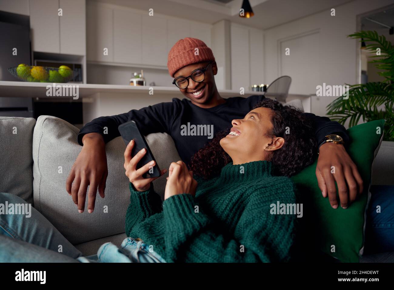 Couple multiethnique se regardant les uns les autres, en riant au sujet du texte sur le téléphone.Sur un canapé dans un appartement moderne Banque D'Images