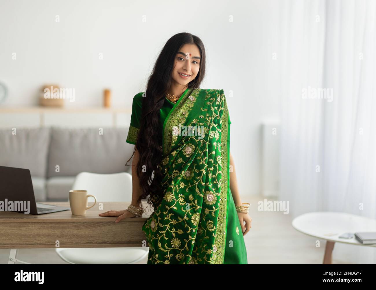 Portrait de la belle dame indienne en robe verte sari s'appuyant sur la table et souriant à l'appareil photo, à l'intérieur Banque D'Images
