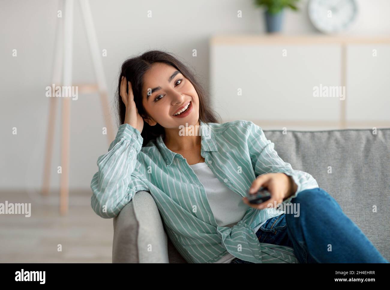 Une femme indienne gaie qui bascule sur les chaînes de télévision avec télécommande et qui regarde la télévision sur le canapé à la maison Banque D'Images