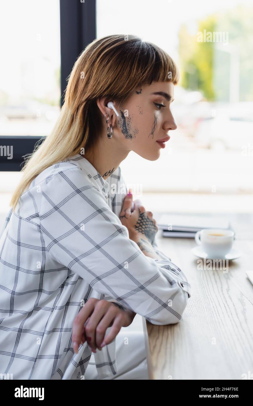 vue latérale d'une femme tatouée dans un écouteur sans fil assis sur le lieu de travail avec des bras croisés Banque D'Images