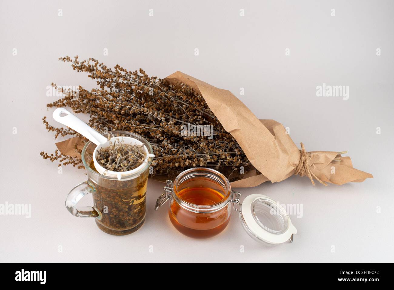 Verre de thé aux herbes, tamis à thé, brindilles au miel et au bois de millepertuis enveloppés de papier brun. Banque D'Images