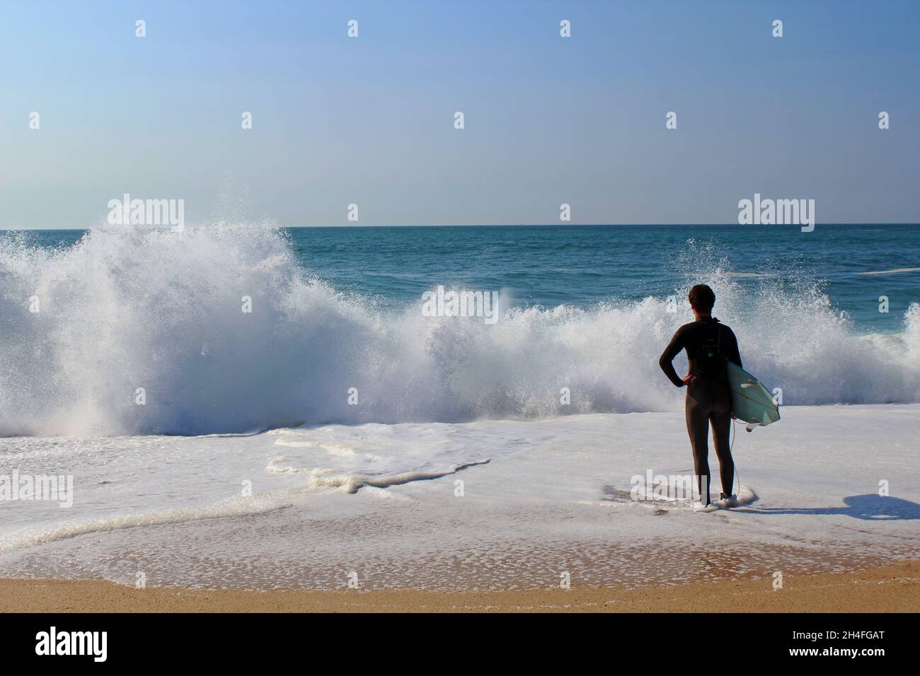 Ein Surfer / Wellenreiter (keine Erkennbarkeit) steht im Neoprenanzug mit seinem Surfbrett unter dem Arm am Strand und schaut auf die Brandungswellen. Banque D'Images