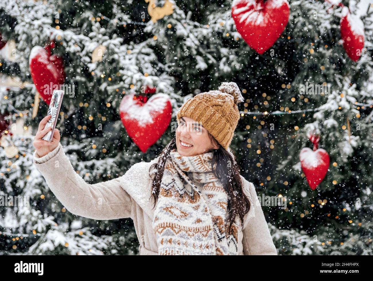 Portrait d'une belle jeune femme heureuse prenant un selfie devant l'arbre de noël le jour de neige en ville Banque D'Images