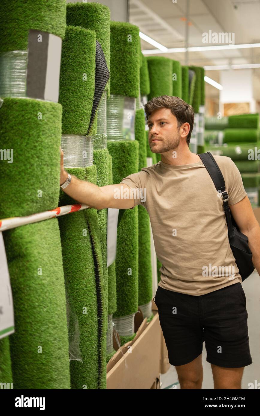 Homme décontracté regardant et touchant un rouleau d'herbe verte artificielle tout en ramassant le matériel pour la décoration de la maison Banque D'Images