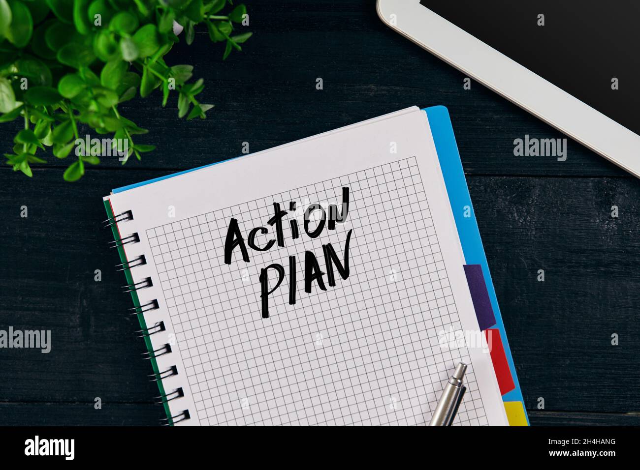 Vue du dessus du carnet ouvert écrit avec inscription AU PLAN D'ACTION. Fleur verte, tablette, stylo et carnet sur fond de bois. Banque D'Images