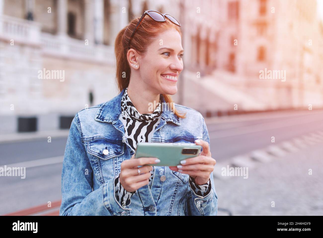 Jeune femme à tête rouge avec un smartphone qui regarde loin dans le coucher du soleil de la ville, à l'extérieur Banque D'Images