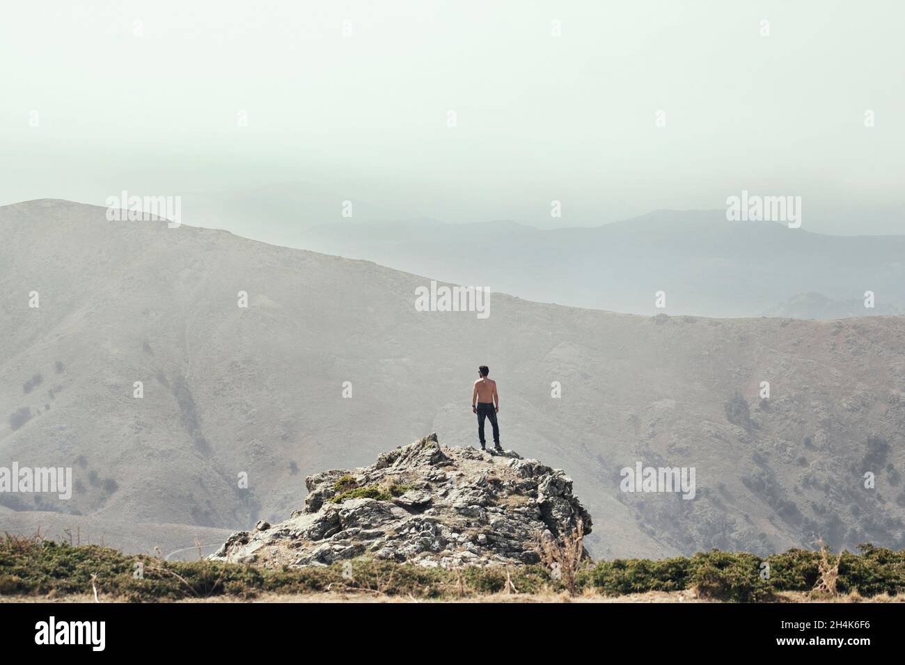 Un jeune homme, vu de l'arrière, se tient au sommet d'un rocher pour regarder la vue des montagnes devant lui. Banque D'Images
