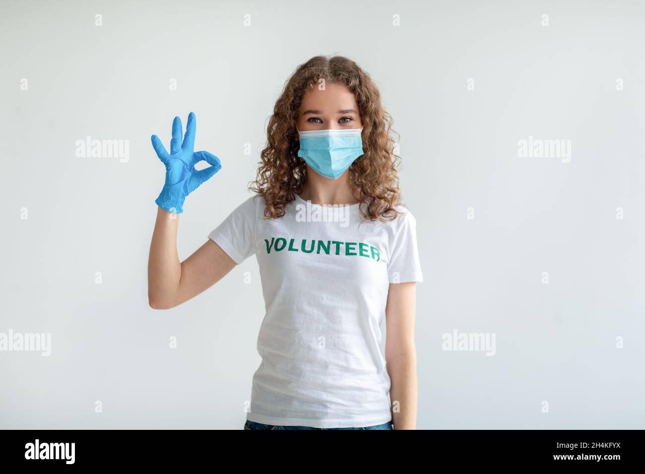 Jeune femme volontaire en t-shirt blanc, masque médical et gants montrant un geste OK et regardant la caméra isolée sur un mur lumineux.Travail libre volontaire, Banque D'Images