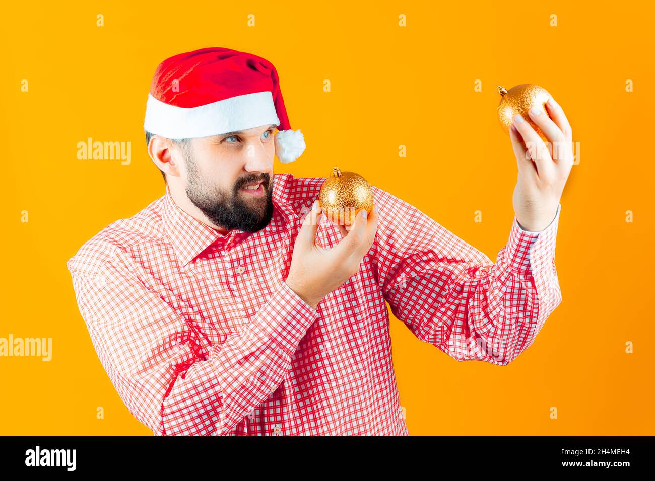 Un homme dans les chapeaux du nouvel an du Père Noël et une chemise à carreaux rouges examine attentivement les jouets du nouvel an - une balle de couleur or. Banque D'Images