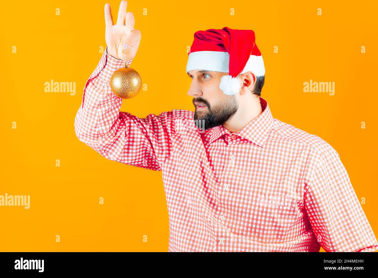 Un homme dans les chapeaux du nouvel an du Père Noël et une chemise à carreaux rouges examine attentivement les jouets du nouvel an - une balle de couleur or. Banque D'Images